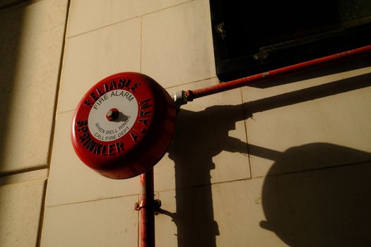 A red fire alarm bell with sprinkler system shadow on exterior wall in Hong Kong.