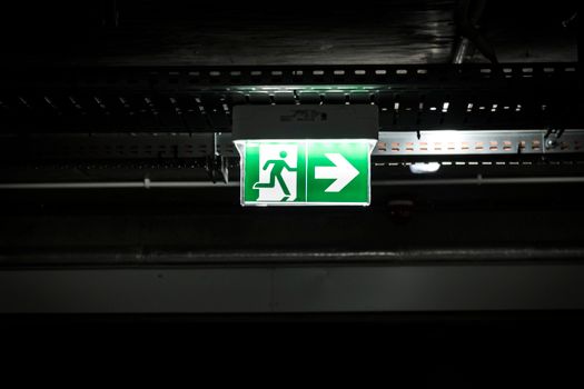 Bright green emergency exit sign with arrow in a dark industrial setting, indicating evacuation route.